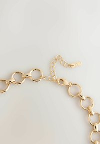 Złoty metalowy łańcuszek na szyję z dużymi, zaokrąglonymi ogniwami i małym regulowanym zapięciem z detalem w postaci metki. Gładka, błyszcząca powierzchnia.