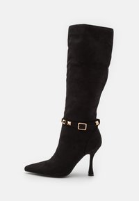 Botas de ante negras hasta la rodilla con puntera afilada, tacón de aguja y una correa decorativa que presenta tachuelas doradas en forma de pirámide y una hebilla cuadrada.