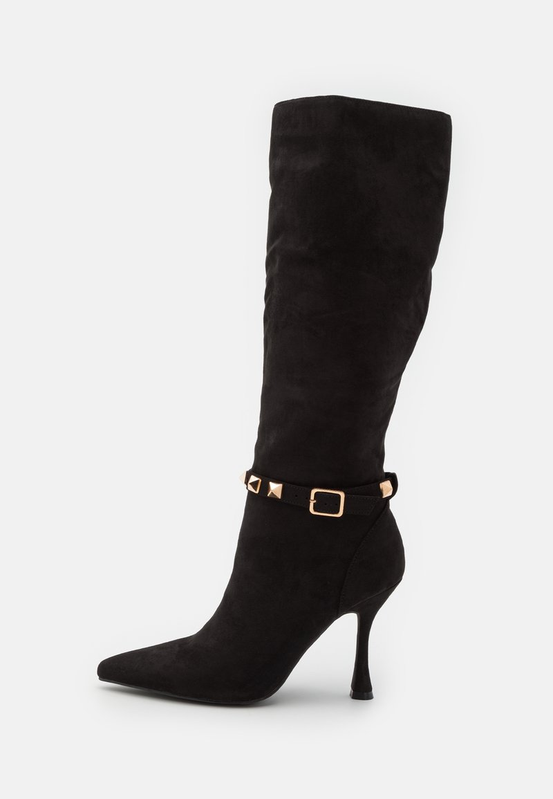 Botas de ante negras hasta la rodilla con puntera afilada, tacón de aguja y una correa decorativa que presenta tachuelas doradas en forma de pirámide y una hebilla cuadrada.