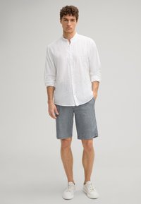 Weißes Leinen-Hemd mit Knöpfen, blaue Shorts und weiße Sneaker. Das Hemd hat einen Stehkragen; die Shorts haben eine subtile Textur und einen mittellangen Schnitt.