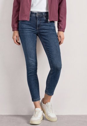 Personne portant un jean slim bleu foncé, des baskets blanches, un haut blanc et une veste zippée bordeaux, debout devant un fond clair uni.