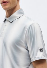 Άνδρας που φοράει ανοιχτό γκρι κοντομάνικη πόλο μπλούζα με γιακά με κουμπιά και μικρό μαύρο λογότυπο στο αριστερό μανίκι.