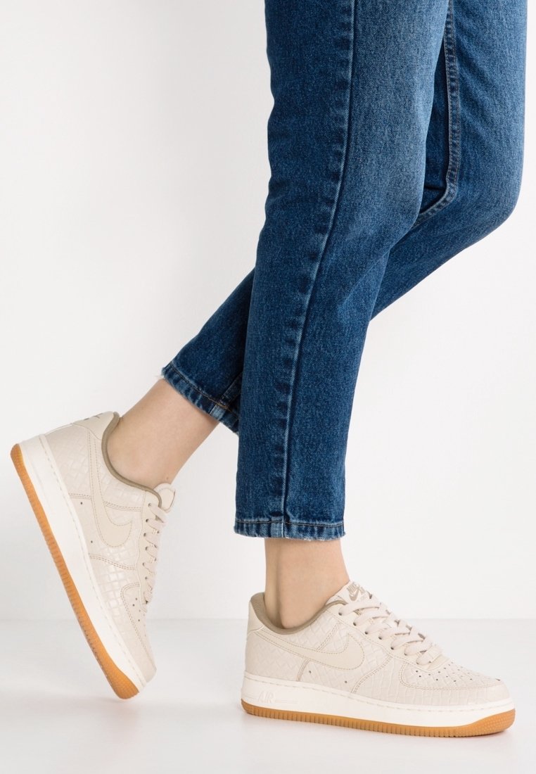 Person som bär blå jeans och beige Nike Air Force 1 sneakers med gummisulor, står framför en vit bakgrund.