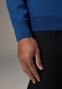 Niebieski sweter wykonany na drutach z ribbowanymi mankietami, wyeksponowany obok ręki spoczywającej na czarnym materiale, podkreślający teksturę i kontrast kolorów.