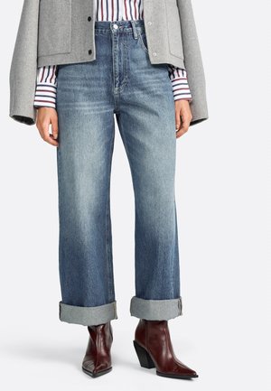 A person wears cuffed, light-wash jeans and stylish burgundy heeled boots, paired with a striped shirt and gray jacket.