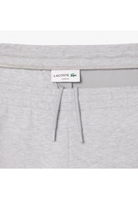 Joggers ajustados de color gris claro con cintura ajustable y una etiqueta de Lacoste cosida en la parte posterior. Material de algodón suave con una textura lisa.
