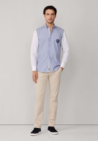 Uomo che indossa pantaloni beige, sneakers neri e una camicia a maniche lunghe con maniche bianche e righe verticali blu, in piedi contro uno sfondo neutro.