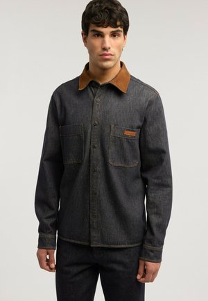 Homme portant une veste en denim foncé avec un col en suède marron et des poches avant, debout devant un fond clair uni.