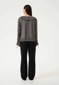 Blusa grigia con colletto plissettato, orlo arricciato e dettagli ai polsini. Modello che indossa pantaloni a gamba larga neri, in piedi su uno sfondo bianco.
