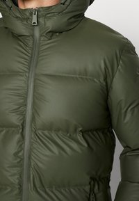 Homme portant une veste matelassée vert olive avec fermeture éclair à l'avant et col montant.