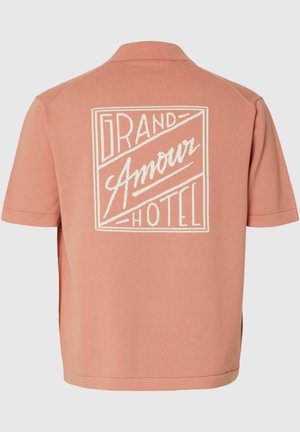 Tył krótkiego, brzoskwiniowego t-shirtu z białym napisem graficznym „Grand Amour Hotel” w prostokątnym obramowaniu.