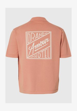 Tył krótkiego, brzoskwiniowego t-shirtu z białym napisem graficznym „Grand Amour Hotel” w prostokątnym obramowaniu.