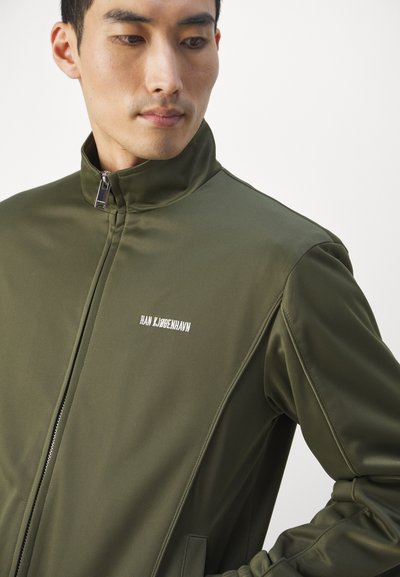 Ελαιόχρωμο σακάκι με ψηλό γιακά, λεία υφή, μπροστινό φερμουάρ και λευκό λογότυπο στην αριστερή πλευρά του στήθους. Κατάλληλο για καθημερινές εμφανίσεις.