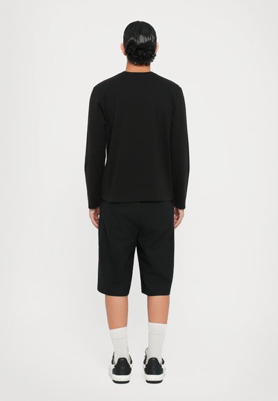 Svart langermet topp kombinert med svarte knelange shorts. Brukt med hvite sokker og svarte joggesko. Enkel, ren design, uten mønstre.
