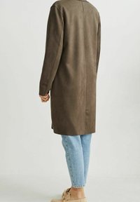 Abrigo largo de ante marrón con corte recto, combinado con jeans de color azul claro y zapatos beige sin cordones. Textura suave y diseño minimalista.