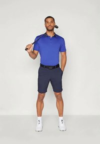 Modra polo majica s ovratnikom, mornarsko modre hlače, črn pas in bele tenisice. Model drži golf palico, stoji proti svetli podlagi.