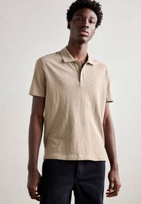Polo shirt a maniche corte beige con patta dei bottoni e texture sottile, abbinato a pantaloni in denim blu scuro. Branding semplice sul petto.