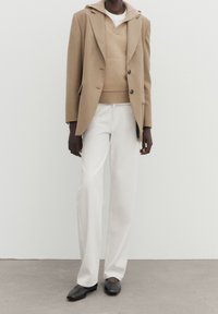 Person iført beige blazer og sweater, hvide bukser og sorte spidse flade sko, stående mod en ensfarvet hvid baggrund.