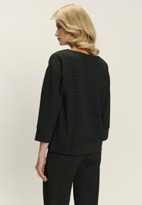 Mujer rubia con cabello ondulado de pie de frente a la izquierda, vistiendo una blusa negra de manga larga con textura y pantalones a juego, contra un fondo liso.