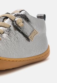 Scarpa in pelle argentata con texture, lacci beige, forma rotonda e suola in gomma. Presenta un'etichetta con il marchio sul lato.