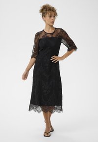 Mujer vistiendo un vestido midi de encaje negro con mangas transparentes, de pie con una mano en la cadera y mirando de lado, sobre un fondo blanco liso.