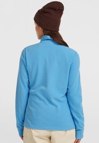 Lysblå fleece-pullover med høj krave og lange ærmer. Blød tekstur; har en afslappet pasform. Bæres sammen med en brun strikket hue.