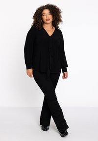 Schwarzes Langarmoberteil mit V-Ausschnitt und drapiertem Vorderteil, kombiniert mit schwarzen Schlaghosen und schwarzen Schuhen; glatte Stoffstruktur.
