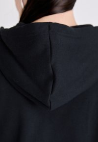 Crna hoodie s mekom teksturom. Ima okruglu kapuljaču i čist dizajn, s vidljivim šavovima duž rubova.