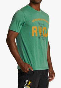Green cotton t-shirt with short sleeves, featuring a large orange RVCA logo and graphic print of a robot arm. Crew neck design, soft texture.