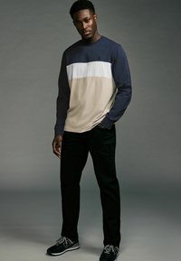Langwijdige shirt in marineblauw, wit en beige horizontale blokken; gecombineerd met zwarte broek en zwarte sportschoenen met grijze accenten.