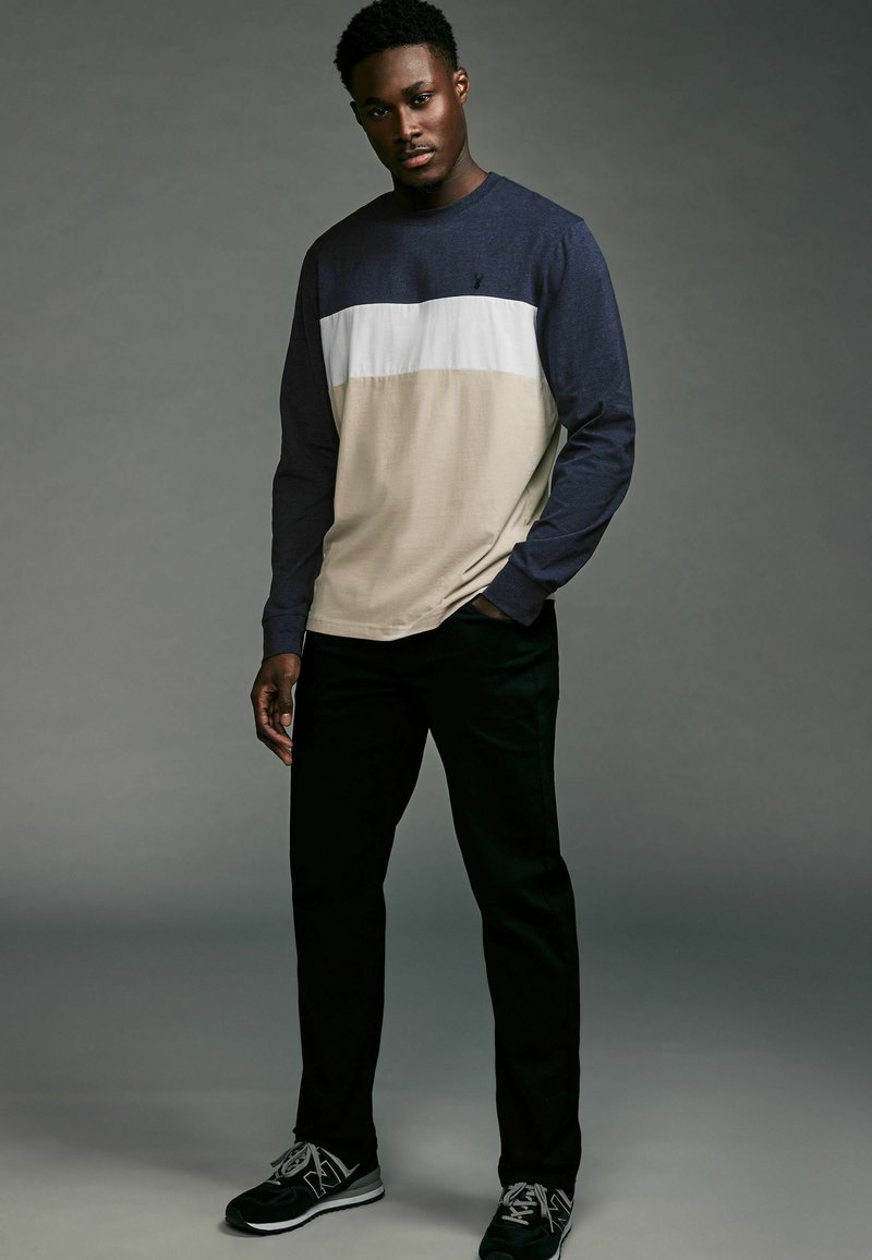 Langwijdige shirt in marineblauw, wit en beige horizontale blokken; gecombineerd met zwarte broek en zwarte sportschoenen met grijze accenten.