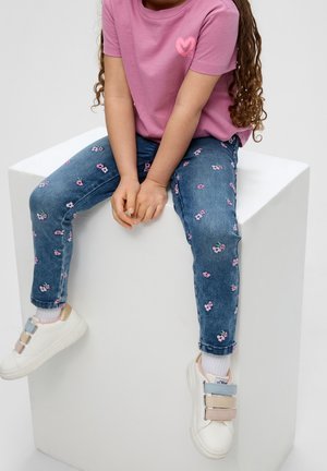 Vaaleanpunainen puuvillapaita, jossa on sydämen kuva, yhdistetty sinisiin farkkuihin, joissa on kukkakirjonta. Valkoiset lenkkarit, joissa on useita nauhavärejä.