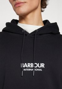 Černá mikina s kapucí z měkké látky, s bílým potiskem loga "BARBOUR INTERNATIONAL" přes hrudník a šňůrkami s kovovými koncovkami.