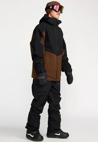 Veste de snowboard noire et marron, dotée d'une fermeture éclair, de grandes poches et d'une capuche. Portée avec un pantalon de neige noir et des gants.