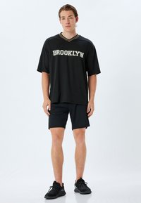 Sort atletisk trøje med "BROOKLYN" i hvid og grå skrift. V-hals design, korte ærmer, parret med sorte shorts og sorte sneakers.