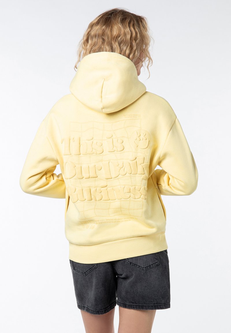 Geel hoodie met een textuur graphic op de achterkant die "Dit is ons donkere bedrijf" leest. Beschikt over een trekkoordcapuchon en geribbelde boorden.