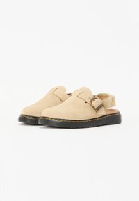 Beige semskede slip-on sko med bakrem, svart gummisåle og oransje sømdetaljer. Har et sirkulært spenneaktsent.