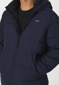 Marineblå pufferjakke med hætte, med en lynlås foran, sidelommer og vandret quiltning. Fremstillet af struktureret, letvægtsstof.