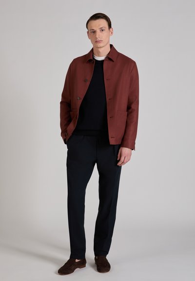 Chaqueta de algodón marrón de corte holgado, con cuello, cierre de botones y dos bolsillos delanteros, combinada con pantalones azul marino y zapatos oscuros.