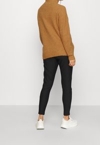 Personne portant un pull à col roulé marron, un pantalon noir à fines rayures et des baskets blanches, debout de dos sur un fond uni.