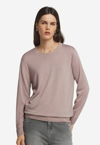 Hellrosa gestrickter Pullover mit rundem Ausschnitt und langen Ärmeln. Weicher Stoff mit gerippten Bündchen und Saum. Über grauen Hosen getragen.