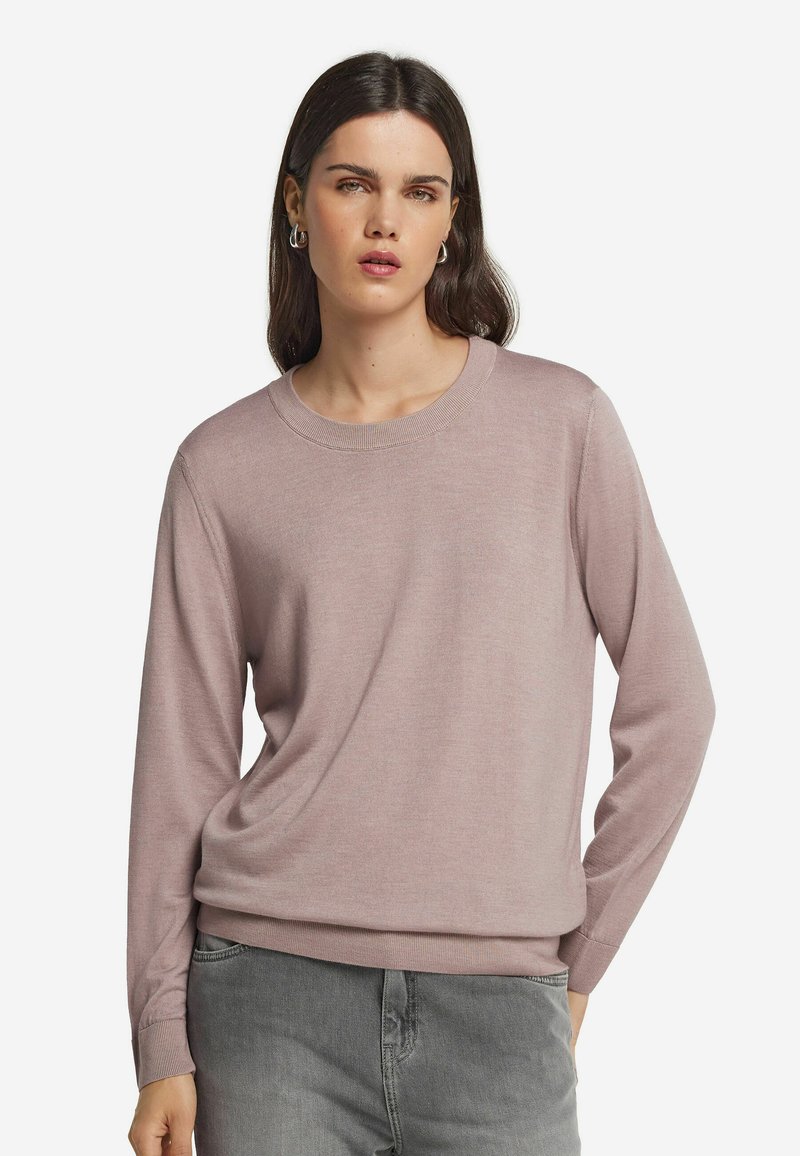 Hellrosa gestrickter Pullover mit rundem Ausschnitt und langen Ärmeln. Weicher Stoff mit gerippten Bündchen und Saum. Über grauen Hosen getragen.