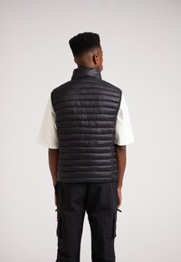 Schwarze gepolsterte Weste mit hohem Kragen, gestepptem Muster und figurbetontem Design. Getragen über einem weißen Kurzarmhemd, von hinten betrachtet.