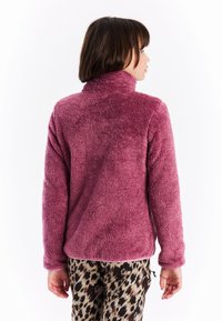 Osoba nosi dugi rukav, visokog vrata ljubičasti flis jaknu i hlače s uzorkom leoparda, okrenuta leđima prema bijeloj pozadini.