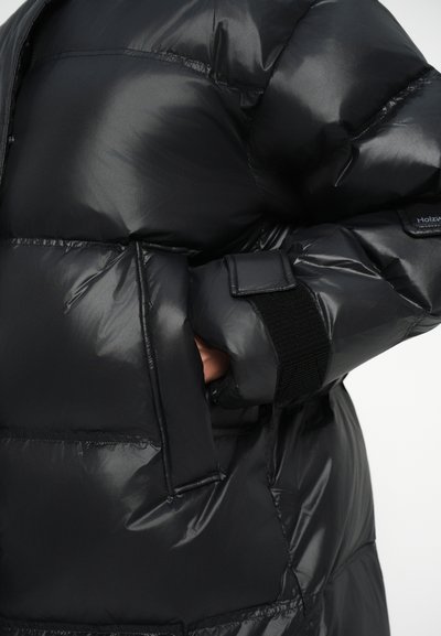 Chaqueta de plumas negra hecha de un material brillante y acolchado. Cuenta con un bolsillo con cremallera y una correa de velcro, además de detalles en los puños elásticos.