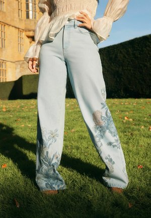 Inimene, kes kannab hele siniseid laia säärega teksapükse lillemustriga tikandiga ja läbipaistvast kreemikast pika varrukaga pluusi, seisab õues rohul.