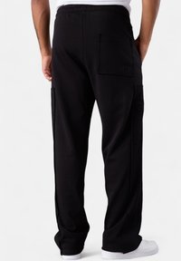 Rückansicht einer Person, die schwarze Cargo-Hosen mit hinteren und seitlichen Taschen sowie weißen Sneakern trägt und vor einem schlichten Hintergrund steht.