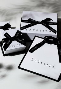 Tre vita lådor med svarta accenter och band, med namnet "LATELITA" i en elegant stil. Den största lådan är fyrkantig.