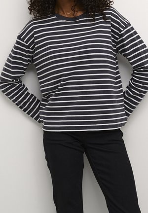 Persoon die een zwart-wit gestreept shirt met lange mouwen en een zwarte broek draagt, staand met handen in de zij tegen een effen achtergrond.