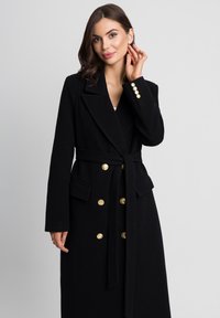 Czarny płaszcz z mieszanki wełny z podwójnymi złotymi guzikami, paskiem do związania oraz kołnierzem z wycięciem. Zawiera funkcjonalne boczne kieszenie.